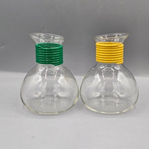 May include: Two clear glass decanters with a bulbous base and angled spout. One has a green coiled neck, the other a yellow coiled neck. The decanters are empty and sit against a gray background.