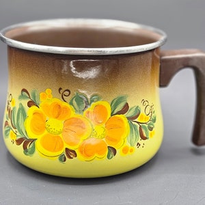Puede incluir: Una taza de esmalte vintage con asa y borde marrón. La taza presenta una base amarilla con un diseño floral en tonos de amarillo, naranja y verde. La taza tiene un degradado de marrón a amarillo.