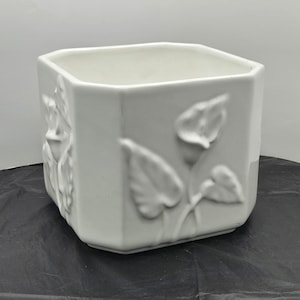 May include: White ceramic planter with a square shape and embossed floral design. The planter has a raised design of leaves and flowers.
