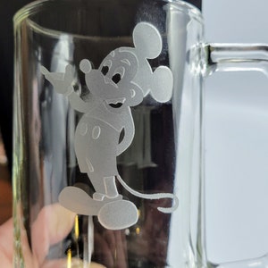 Puede incluir: Una taza de vidrio transparente con un diseño grabado en blanco de Mickey Mouse. Mickey está de pie con la mano derecha levantada y la mano izquierda en la cadera.