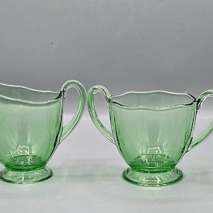 Könnte beinhalten: Ein Paar grüne Vintage-Servierschalen aus Glas, eine Sahnekännchen und eine Zuckerdose. Das Sahnekännchen hat einen Ausguss und einen Griff. Die Zuckerdose hat zwei Griffe und einen gewellten Rand. Beide stehen auf einem runden Sockel.