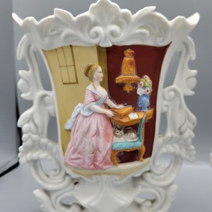 Puede incluir: Jarrón de porcelana blanca con un diseño ornamentado en relieve. El jarrón representa una escena de una mujer con un vestido rosa de pie en un escritorio con un gato sentado en una silla. La escena está enmarcada por un borde decorativo.