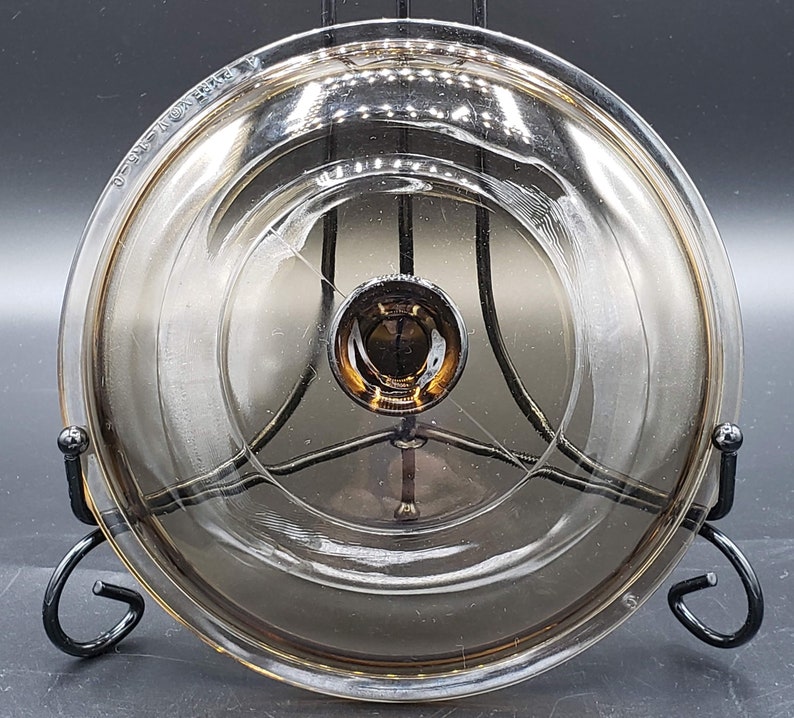 May include: A clear glass lid with a brown tint and a metal knob. The lid is on a black metal stand.