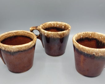 May include: Three brown and white glazed ceramic mugs with a speckled design. The mugs have a rounded shape and a single handle.