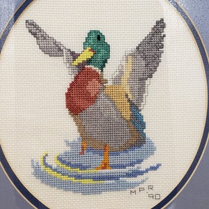 May include: A framed cross-stitch of a mallard duck with green head and wings spread, standing on a blue and yellow water surface. The cross-stitch is signed "M.P.R. '90".
