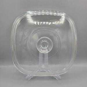 May include: A clear, square glass lid with rounded corners. The lid features a central handle and a slightly raised rim. The glass is transparent, allowing a view of the surface below. The lid is designed for cookware.