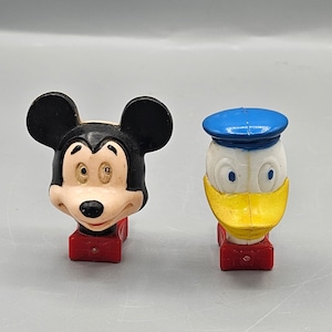 May include: Two vintage plastic character head figurines. Mickey Mouse is black with large ears and a red base. Donald Duck is white with a yellow beak and blue sailor hat, and a red base.