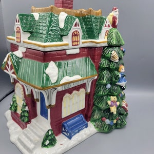 Puede incluir: Un tarro de galletas de Navidad de cerámica con forma de estación de tren con un techo verde y un árbol de Navidad con adornos y un oso de peluche. El tarro dice "Train Station" en la parte delantera.