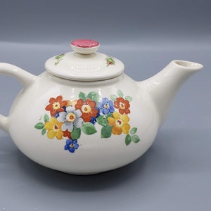 May include: A white ceramic teapot with a floral design. The teapot has a pink lid and a spout. The flowers are red, orange, yellow, blue, and green.