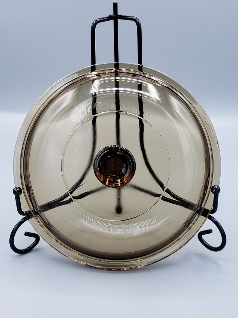 May include: A clear glass lid with a brown tint, resting on a black metal stand. The lid has a small knob in the center.
