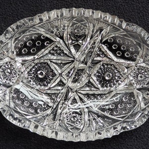 May include: Clear, oval-shaped crystal dish with intricate cut glass patterns. The dish features a scalloped edge and a detailed design of stars, flowers, and geometric shapes. A decorative piece for serving or display.