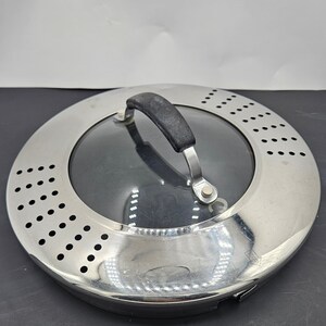 Puede incluir: Una tapa redonda de plata y vidrio con un asa negra. La tapa tiene un borde perforado. La tapa está diseñada para usarse con una olla o sartén.