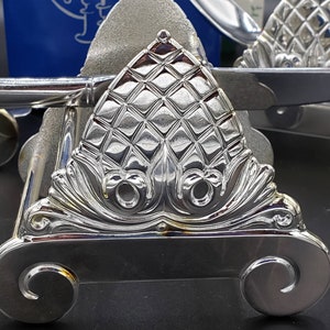 Puede incluir: Un porta servilletas decorativo plateado con un diseño de piña. El porta servilletas tiene un diseño de rollo y dos pies redondeados.