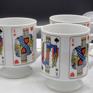 Puede incluir: Conjunto de tres tazas de cerámica blanca con ribete dorado. Cada taza presenta un diseño de carta de juego diferente con el Rey de Picas, el Rey de Corazones y el Rey de Tréboles.
