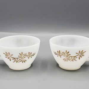 Op de afbeelding: Twee witte keramische theekopjes met een gouden bloemenmotief. De kopjes hebben een afgeronde vorm en een handvat aan de rechterkant.
