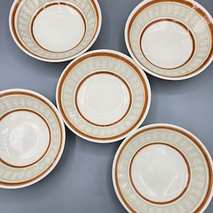 Puede incluir: Cinco cuencos de cerámica blanca con ribete de patrones marrones y verdes. Los cuencos están dispuestos en círculo sobre una superficie gris.