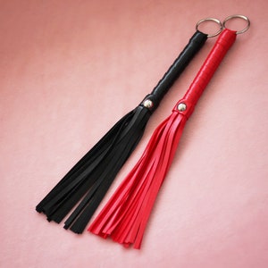 Puede incluir: Dos l&aacute;tigos de cuero con borlas, uno negro y otro rojo, sobre un fondo rosa.