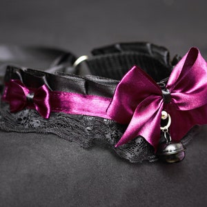 May include: A black choker with a purple satin bow and a bell charm. The choker is made of lace and satin.