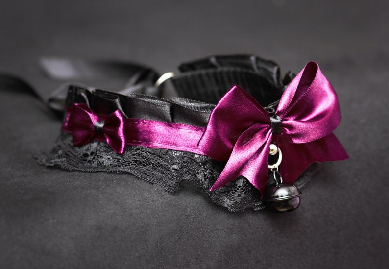 May include: A black choker with a purple satin bow and a bell charm. The choker is made of lace and satin.