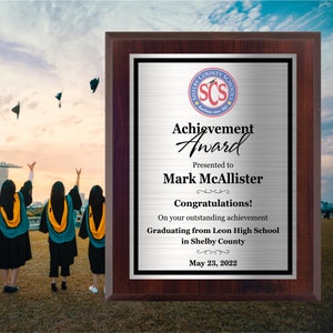 May include: A rectangular wooden plaque with a brushed silver metal plate featuring the Shelby County Schools logo, the text Achievement Award, Mark McAllister, Congratulations, and details about graduating from Leon High School on May 23, 2022.