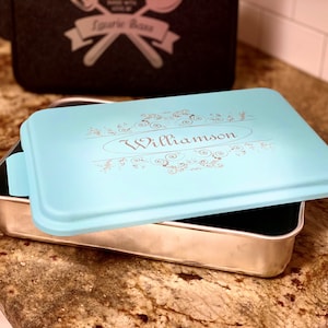 May include: A rectangular baking pan with a light blue lid. The lid is engraved with the name "Williamson" in an elegant script, surrounded by decorative flourishes. A black bag with text is in the background.