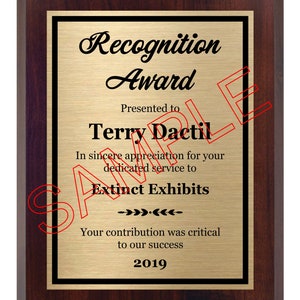 May include: A mahogany-colored plaque with a gold-colored plate. The plaque has a black border and the text "Recognition Award" in a black cursive font. The text "Presented to Terry Dactil" is in black text. The text "In sincere appreciation for your dedicated service to Extinct Exhibits" is in black text. The text "Your contribution was critical to our success" is in black text. The text "2019" is in black text.