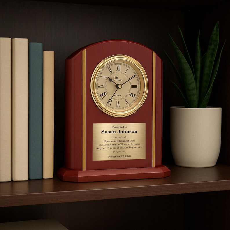 May include: A wooden desk clock with a gold-colored clock face and Roman numerals. The clock has a rectangular base and a curved top. A gold plaque reads "Presented to Susan Johnson" and includes text about retirement. A potted plant and books are in the background.