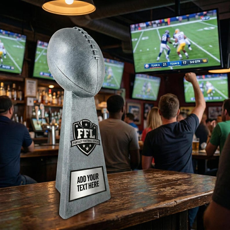 May include: A gray football-shaped trophy with a base featuring the text "FFL FANTASY FOOTBALL LEAGUE" and a space to add text. The trophy is on a wooden bar, with multiple televisions showing a football game in the background.