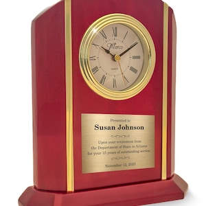 May include: A burgundy-colored desk clock with a gold-tone clock face and trim. The clock face has Roman numerals and the brand name "Marco". A gold plaque reads "Presented to Susan Johnson" and includes a retirement message dated November 12, 2023.