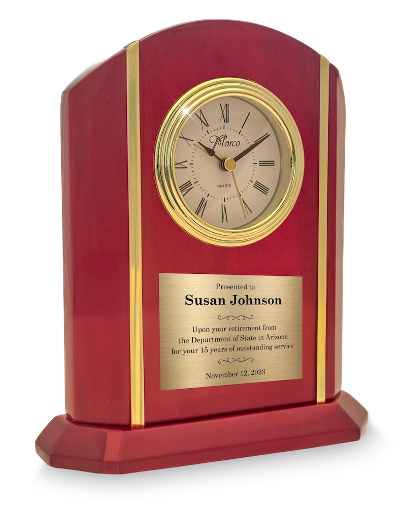 May include: A burgundy-colored desk clock with a gold-tone clock face and trim. The clock face has Roman numerals and the brand name "Marco". A gold plaque reads "Presented to Susan Johnson" and includes a retirement message dated November 12, 2023.