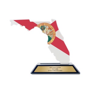 May include: A clear acrylic trophy shaped like the state of Florida with the Florida state flag printed on it. The trophy sits on a black base with a gold plate that says "Add your custom text here. Up to 3 lines included."