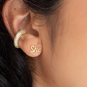 May include: A close-up of a person's ear with a gold ear cuff and a small gold earring with the word "She" in cursive.