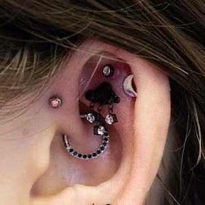 Peut inclure: Une oreille percée à plusieurs endroits, ornée de divers bijoux. On y trouve un clou d'oreille en forme d'araignée noire, un charm nuage noir avec des cristaux pendants, un croissant de lune et une boucle d'oreille noire. Les bijoux se détachent sur la peau.