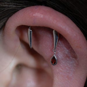 Pode incluir: Close-up de uma orelha com vários piercings. Um piercing apresenta uma barra de prata com uma pedra preciosa vermelha em forma de gota pendurada. Outro piercing é uma barra de prata. Vários outros piercings são visíveis com pequenos pregos de prata.