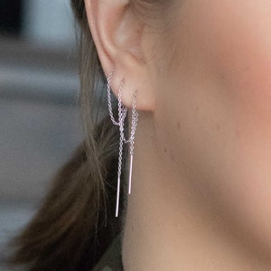 May include: A close-up of a person's ear with three silver chain earrings. The earrings are dangling and have a delicate, minimalist design.