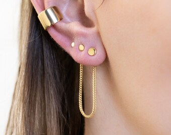 Puede incluir: Primer plano de un brazalete de oreja dorado y tres pendientes de botón dorados en una oreja. Un pendiente de cadena dorada cuelga del pendiente inferior. La joyería es brillante y reflectante, sobre un fondo borroso.