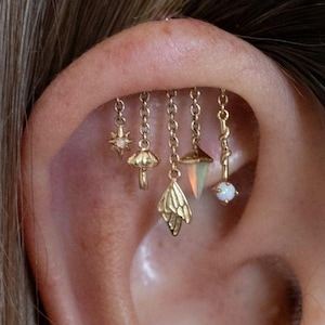 Könnte beinhalten: Goldfarbener Ohrschmuck mit einer Reihe von baumelnden Anhängern. Die Anhänger umfassen einen Stern, einen Pilz, ein Blatt und eine geometrische Form, die jeweils an zarten Ketten hängen. Ein kleiner opalartiger Stein ist ebenfalls enthalten. Der Schmuck wird am Ohr getragen.