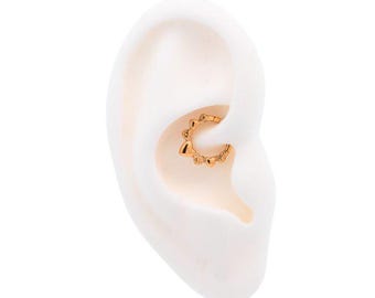 Pode incluir: Um anel de septo dourado com um design decorativo. A joia é colocada em um modelo de orelha branco, mostrando os detalhes intrincados e a forma do anel. O anel apresenta pequenos enfeites espaçados.