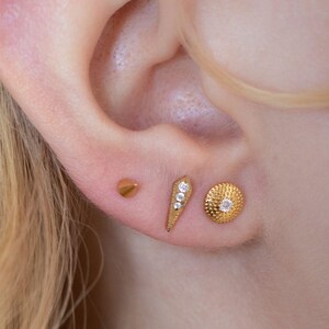 May include: Close-up of three gold earrings on an ear. The earrings include a small cone stud, a triangular earring with three small clear stones, and a round earring with a textured surface and a single clear stone. The earrings are gold in colour.