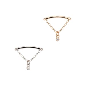 Puede incluir: Dos delicadas piezas de joyería. Una es dorada, la otra plateada. Cada una presenta una barra curva conectada a una cadena que desciende hasta una pequeña gema redonda y transparente. La joyería está sobre un fondo blanco.