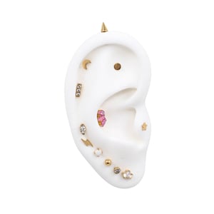 Puede incluir: Una colección de pendientes dorados, que incluyen tachuelas, aros y una espiga, exhibidos en un modelo de oreja blanca. Los pendientes presentan varios diseños, como una luna, una estrella, un rayo y detalles de piedras preciosas. Los pendientes son pequeños y delicados.
