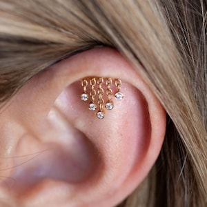 Könnte beinhalten: Ein goldfarbener Ohrschmuck mit feinen Ketten, an denen jeweils ein kleiner, klarer Edelstein hängt. Der Schmuck ist für den oberen Ohrknorpel gedacht und verleiht dem Ohr einen dezenten, aber auffälligen Glanz.