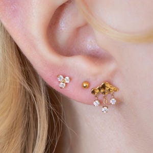 Peut inclure: Gros plan d'une oreille ornée de trois boucles d'oreilles dorées. L'une est un groupe de trois pierres claires, une autre une petite boule dorée, et la troisième un nuage avec trois pierres claires pendantes.