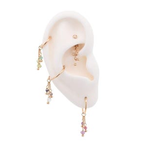 May include: Gold-coloured ear piercings with dangling charms featuring colourful gemstones. The ear includes multiple hoop earrings and a chain with small, dangling beads. The gemstones are in shades of green, purple, pink, and white.