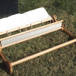 Peut inclure: Un métier à tisser à peigne envergeur en bois avec un cadre de couleur claire, des boutons de réglage latéraux, un peigne central en métal à fentes verticales fines et une chaîne en tissu blanc fixée au rouleau arrière par des cordons.