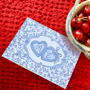 May include: A greeting card with blue floral design and two heart-shaped lockets with the letters "M" and "Z" inside. A bowl filled with fresh, red cherries sits next to the card. The background is a textured red surface.