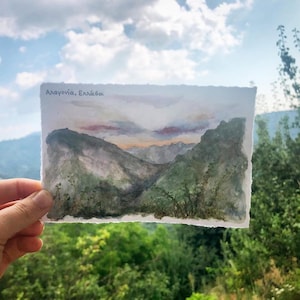 May include: Watercolor painting of a mountain landscape with green and gray tones, under a cloudy sky. The painting has the text "Αλαγονία, Ελλάδα" at the top. The artwork is held in front of a background of green foliage.