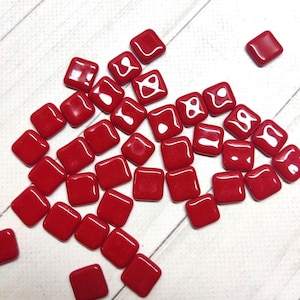 May include: A collection of small, square, glossy red tiles scattered on a light-colored wooden surface. The tiles have rounded corners and a smooth, reflective finish, suggesting they are made of glass or a similar material. They are likely for mosaic art.