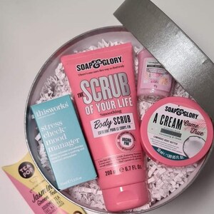 Pu&ograve; includere: Una scatola regalo rosa piena di prodotti per il bagno e il corpo. La scatola contiene un tubo di scrub corpo rosa con il testo "The Scrub of Your Life" e "Smoothing Body Scrub", una piccola bottiglia blu con il testo "thisworks stress check mood manager", un piccolo barattolo rosa con il testo "SOAP & GLORY A Cream Come True", e una piccola bottiglia rosa con il testo "VANILLA BEAN SOAK & SLEEP"