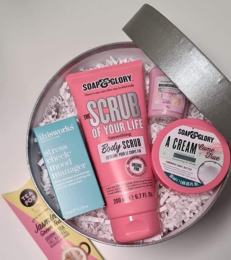 Pu&ograve; includere: Una scatola regalo rosa piena di prodotti per il bagno e il corpo. La scatola contiene un tubo di scrub corpo rosa con il testo "The Scrub of Your Life" e "Smoothing Body Scrub", una piccola bottiglia blu con il testo "thisworks stress check mood manager", un piccolo barattolo rosa con il testo "SOAP & GLORY A Cream Come True", e una piccola bottiglia rosa con il testo "VANILLA BEAN SOAK & SLEEP"